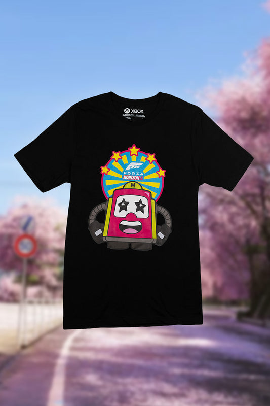 A black short-sleeve T-shirt with a colorful cartoon arcade machine graphic on the front, featuring an Xbox logo printed inside the collar. The shirt is displayed against a blurred background of a road and pink cherry blossom trees under a blue sky.